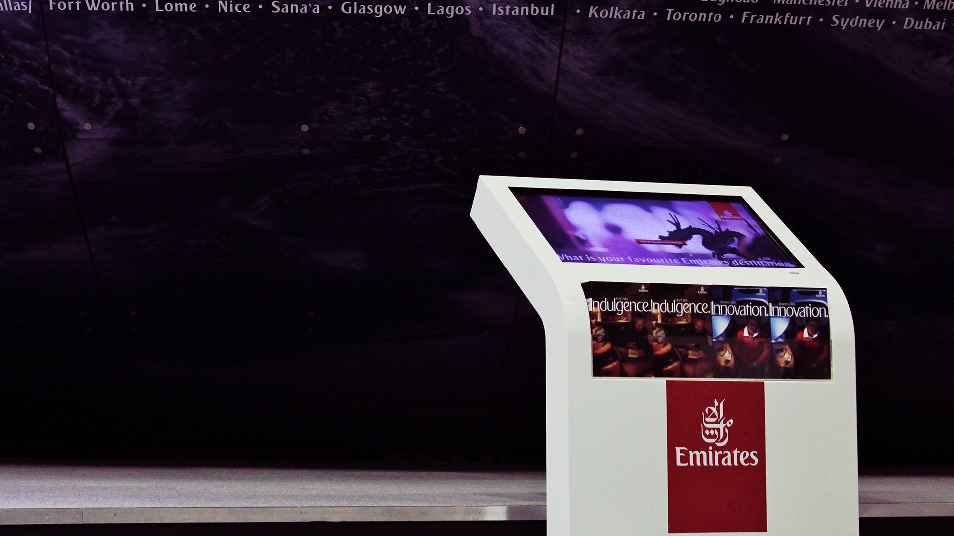 A freestanding Emirates touchscreen kiosk against the exhibition's illuminated world map.