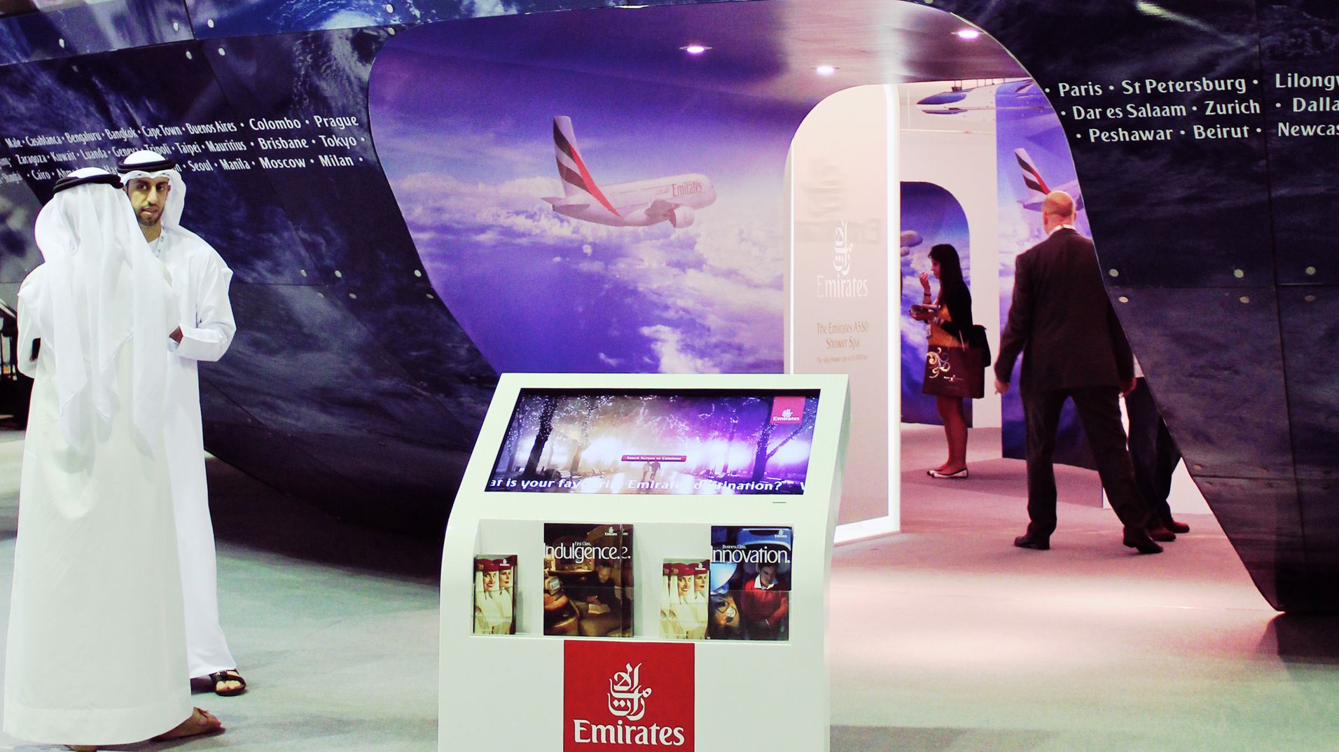 Visitors passing an Emirates kiosk and curved destination display at the exhibition.