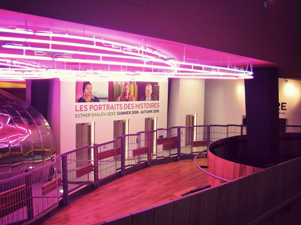 A curved gallery walkway illuminated in pink beneath a display of visitor portraits.