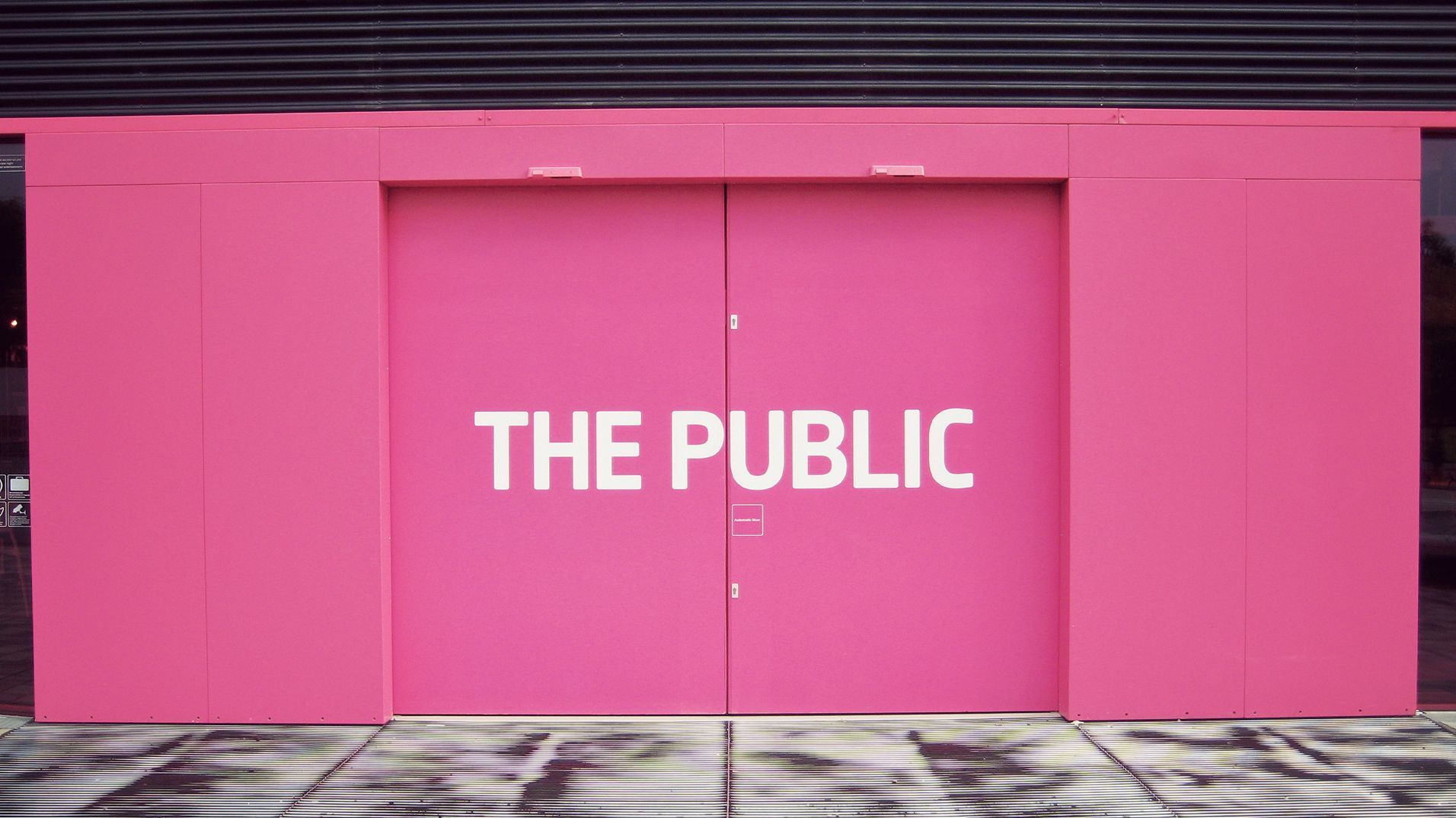The Public entrance illuminated pink with its name projected across the doors.